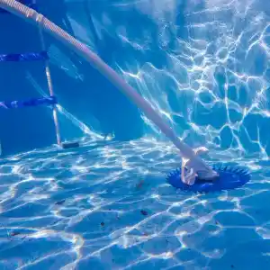 19007-2 | PisciManía Underwater view in a blue pool: a pool-cleaning vacuum head on the floor attached to a long pole, with a metal ladder nearby and sunlight ripples on the pool surface.