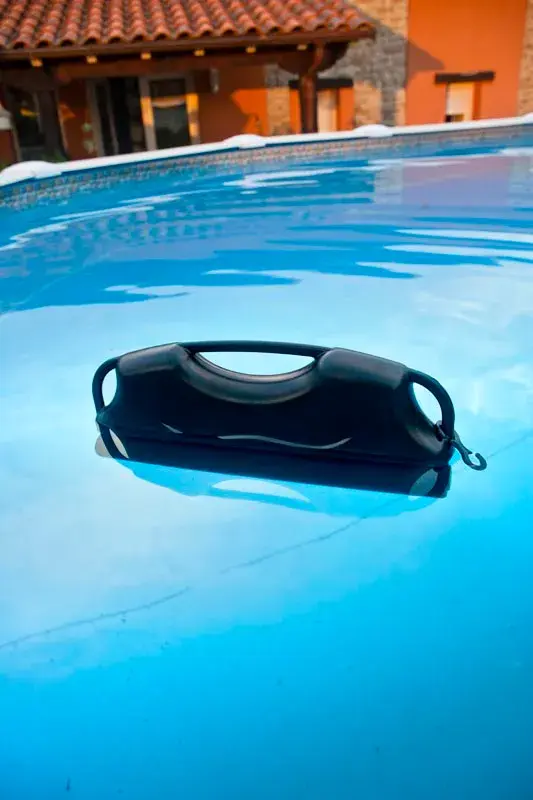 40580-2 | PisciManía Black pool flotation device with handles drifting on the surface of a bright blue pool, orange building in the background.
