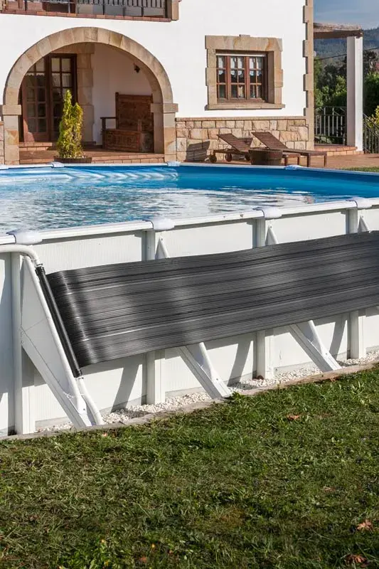 ar2069-1 | PisciManía Above-ground pool with a black solar cover rolled on a white frame, near a stone-clad house with arches and lounge chairs on the patio.
