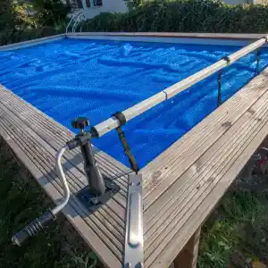 enrollador-maximo-550-cm-de-gre-1 | PisciManía Above-ground pool on a wooden deck with a blue mesh cover and a manual reel system for rolling the cover, including a crank handle and straps attached to the railing.