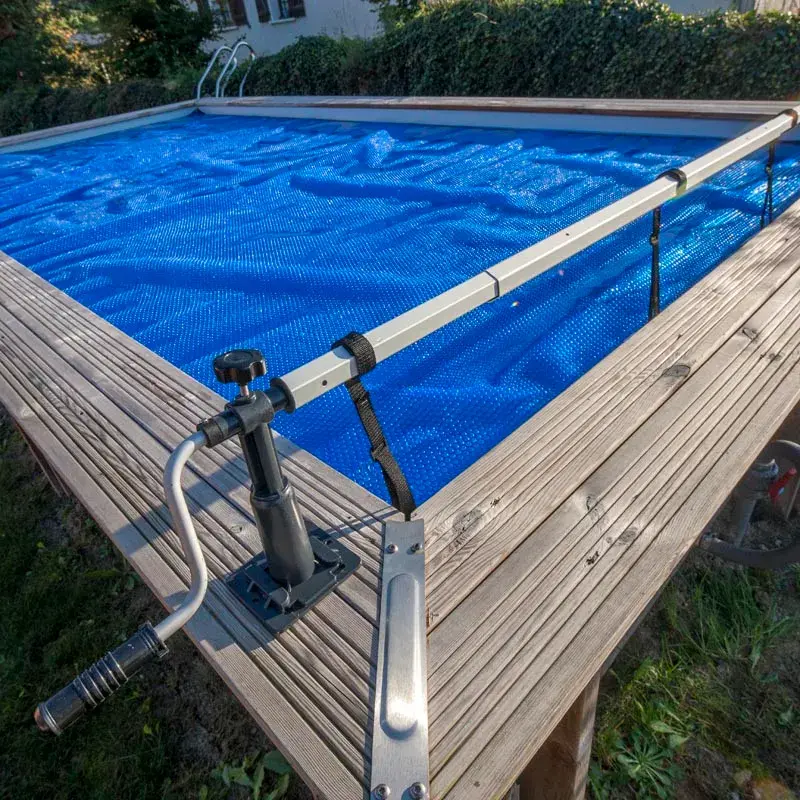 enrollador-maximo-550-cm-de-gre-1 | PisciManía Above-ground pool on a wooden deck with a blue mesh cover and a manual reel system for rolling the cover, including a crank handle and straps attached to the railing.