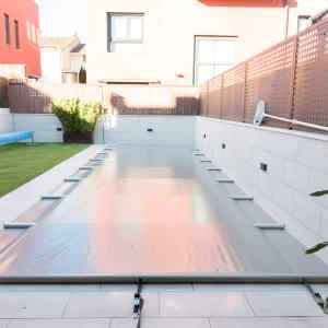 Rectangular pool in a modern courtyard covered by a transparent pool cover, with tiled patio and brown lattice fence.