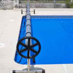 Manual pool cover roller along the edge of a rectangular blue pool, with a large black hand wheel in the foreground.